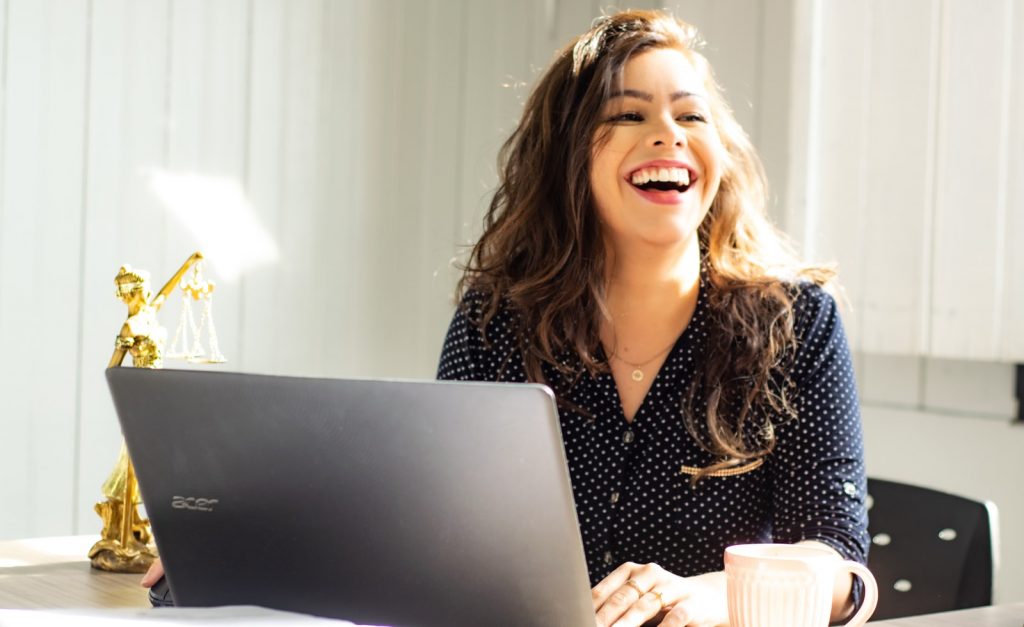A woman laughs while sitting at a desk with a laptop and a pink mug, next to a small statue of Lady Justice, embodying an environment that promotes wellbeing.