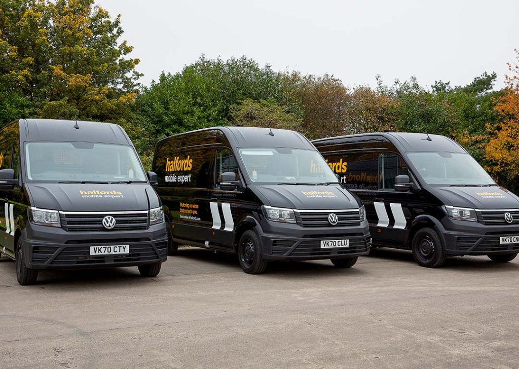 Three black vans with "Halfords Mobile Expert" branding are parked in a row on a concrete surface, ensuring safety and wellbeing, with trees in the background.
