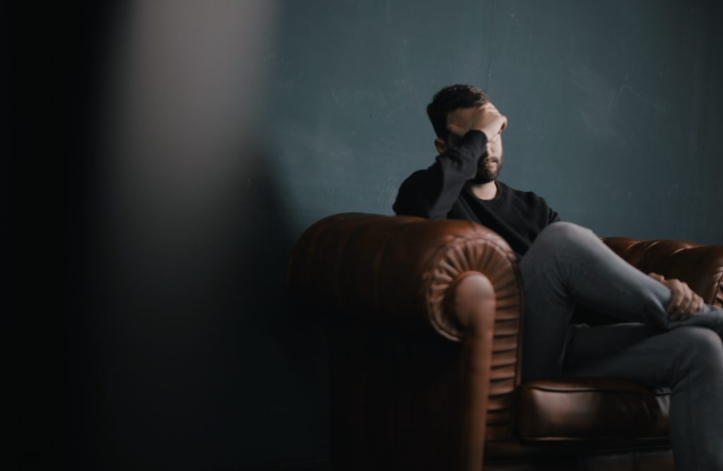 A person sits on a brown leather chair with a hand covering their face, against a dark green wall, contemplating their wellbeing.