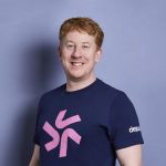 A person with short red hair is smiling and wearing a navy blue shirt with a pink symbol and "deputy" printed on the sleeve, standing against a light purple background, radiating an aura of wellbeing.