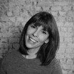 A person with shoulder-length hair and bangs is standing in front of a brick wall, smiling. They are wearing a plain shirt, radiating an aura of wellbeing. The photo is in black and white.