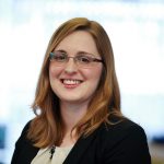 A person with long, light brown hair and glasses, wearing a black blazer and a light-colored top, smiles at the camera against a blurred background, radiating an aura of wellbeing.