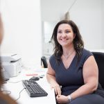 A woman in a black dress sits at a desk, smiling at someone off-camera. A computer keyboard and office equipment are visible on the desk in front of her, reflecting an environment that prioritizes wellbeing.