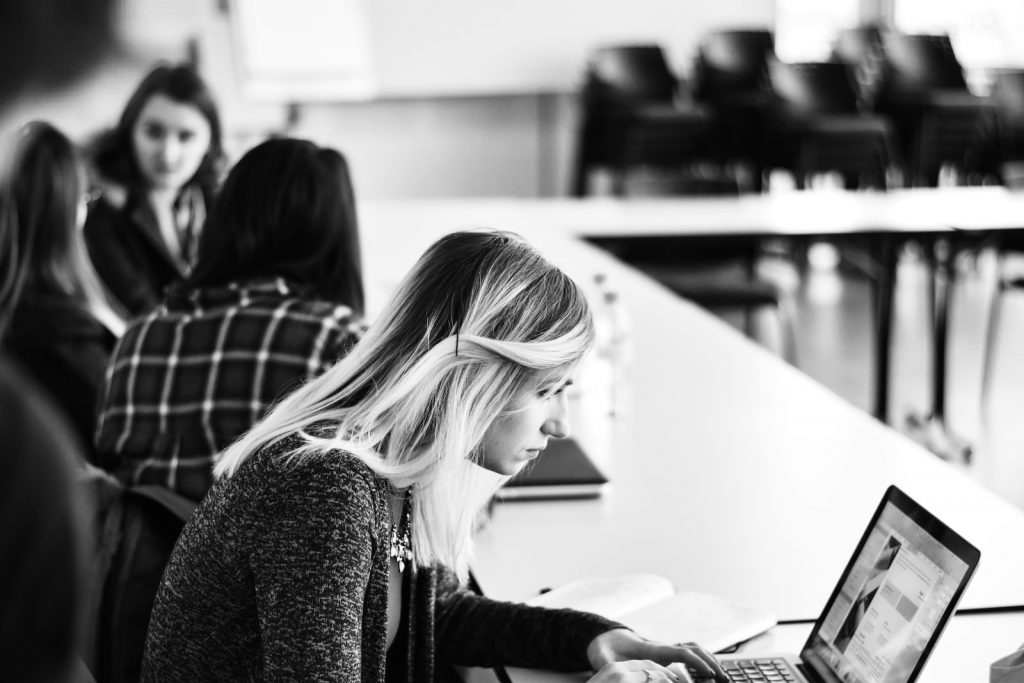 Black and white image of a group of people working at tables, with a woman in the foreground focused on a laptop, ensuring her wellbeing.
