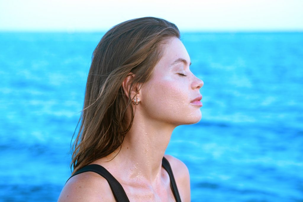 A woman with closed eyes, damp hair, and glistening skin stands in front of the ocean, radiating a sense of wellbeing.