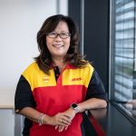 A woman wearing a DHL uniform smiles while standing next to a window, radiating an aura of wellbeing and safety.