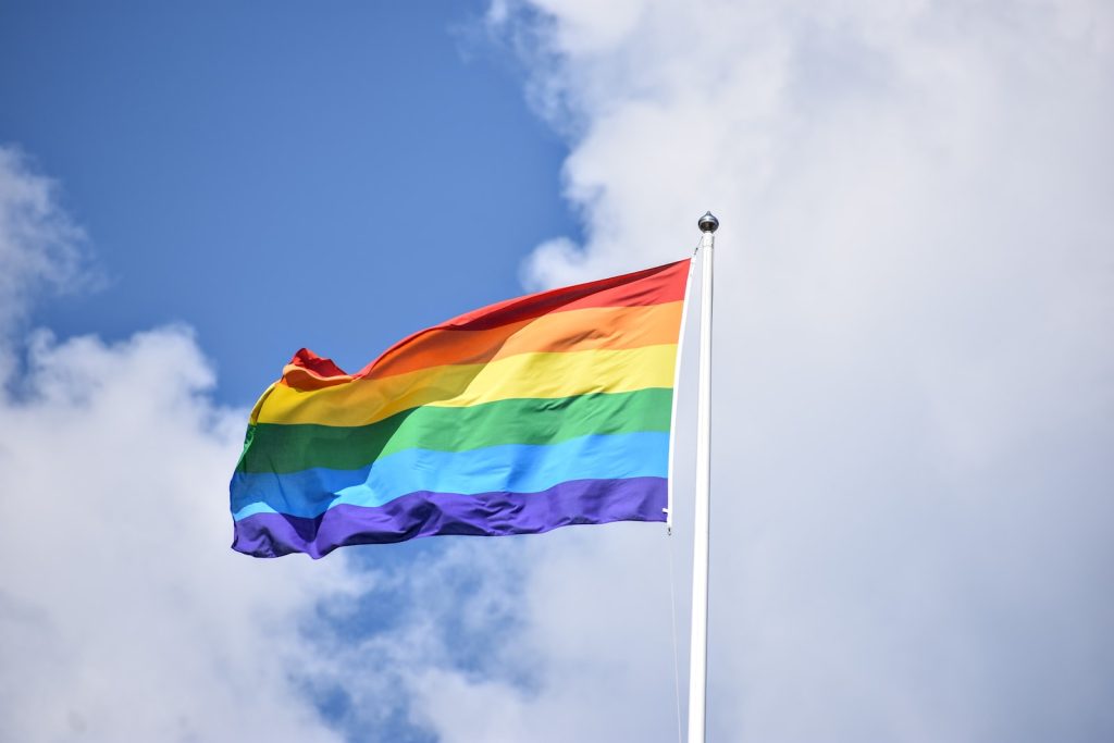 A rainbow flag waves on a flagpole, promoting wellbeing and safety, against a backdrop of a blue sky with scattered clouds.