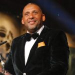 A man in a black velvet tuxedo and bow tie smiles while speaking at a podium, emphasizing the importance of wellbeing and safety.