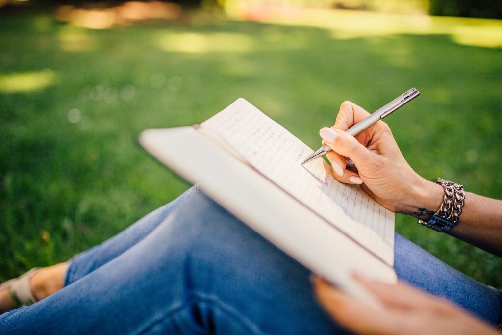 A person sits on grass, writing in a notebook with a pen, finding solace in the peaceful environment that promotes their wellbeing.