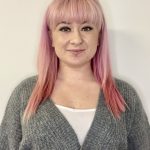 A person with long pink hair and a lip piercing, promoting wellbeing, is wearing a grey cardigan and white top, standing against a plain white background.