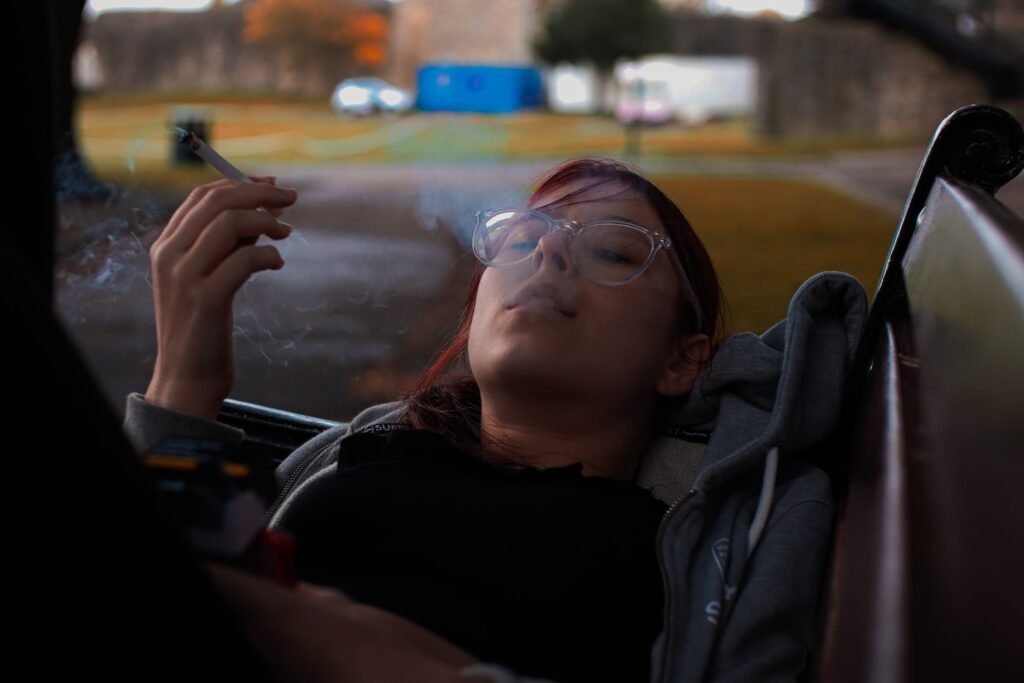 A person with glasses and dyed hair is lying on a bench outdoors, smoking a cigarette. The background features blurred greenery and structures, creating a serene setting. However, it's important to consider health and wellbeing while enjoying the outdoors.