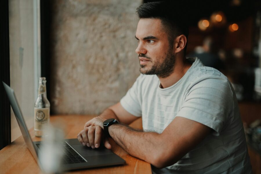 man in white crew neck t-shirt sitting by the table using laptop computer