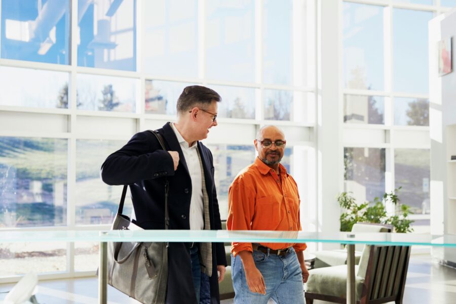 Two men walking and conversing inside a brightly lit modern building with large windows, ensuring their wellbeing. One carries a bag over his shoulder.