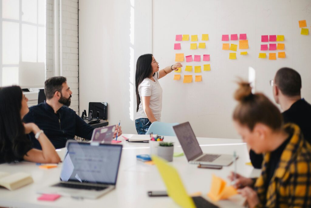 A woman stands at a whiteboard covered in sticky notes, presenting an innovative health plan to three seated individuals. Two open laptops are visible on the table, emphasizing the importance of digital tools for enhancing wellbeing.