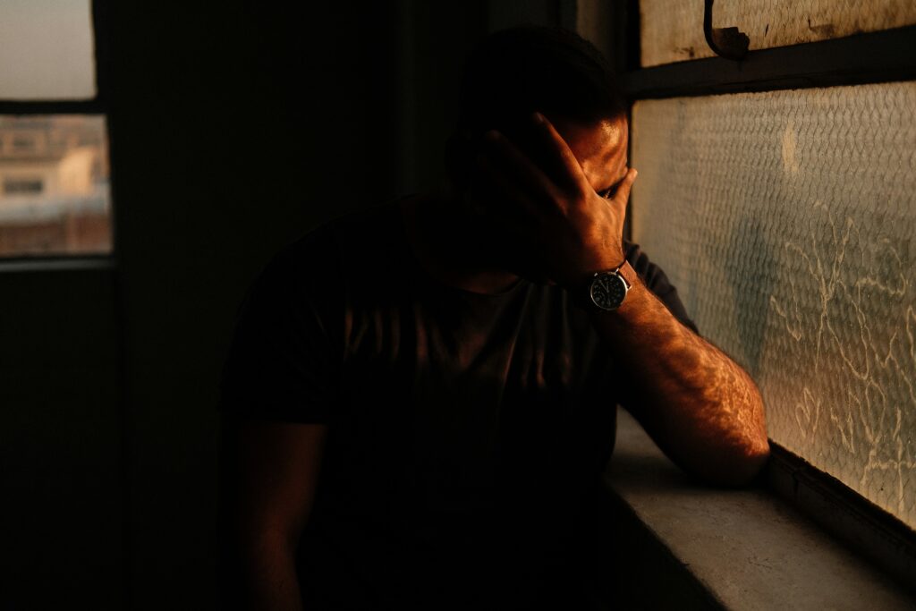 A person stands by a window, covering their face with one hand, pondering their job responsibilities while wearing a watch in the dim light.