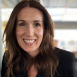 Angela Moyle, a woman with long brown hair, smiles warmly while wearing a dark blazer and necklace, standing indoors with a blurred background.