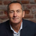 Bradley Honnor, in a navy blazer and striped shirt, sits with hands clasped on a wooden chair against a brick wall background.