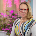 Claudia Nicholls, wearing red glasses and a colorful striped top, sits gracefully in front of a blurred background adorned with pink flowers.