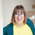 Debbie Hancock, with short hair and glasses, smiles warmly while wearing a yellow shirt and green jacket, standing near a wooden staircase.