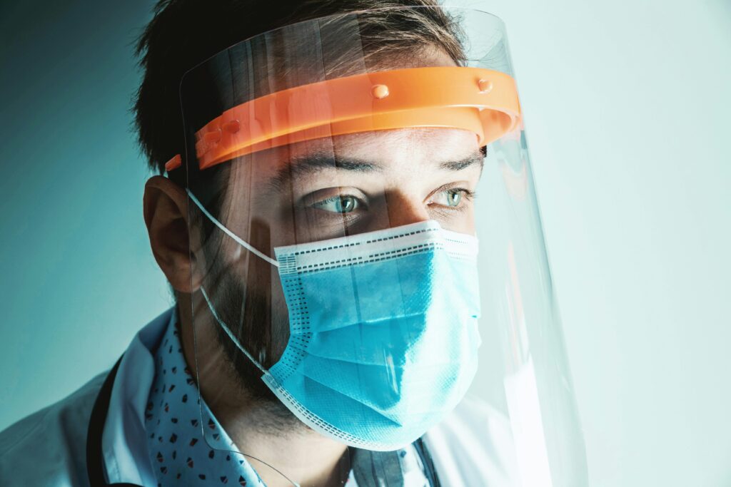 A person, possibly covered by private medical insurance, wearing a protective face shield, blue surgical mask, and stethoscope, looks to the side with a focused expression.
