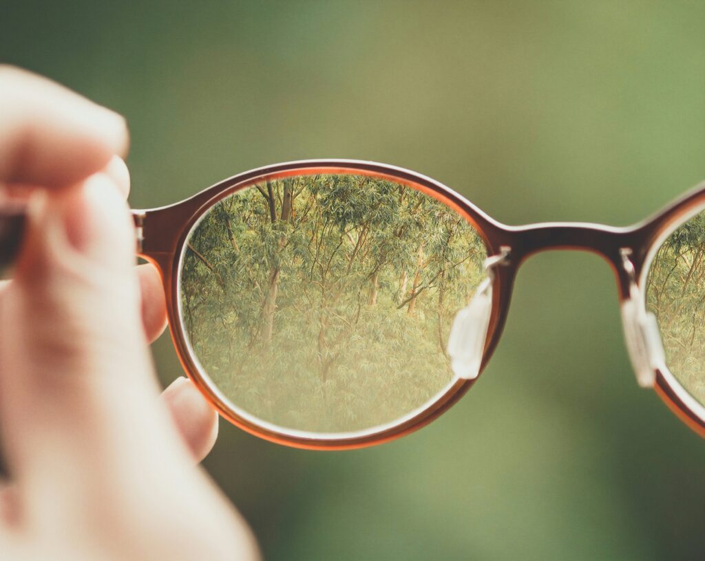 A hand holds a pair of glasses, enhancing visual accessibility by focusing on a clear view of trees through the lenses, with a blurred green background.