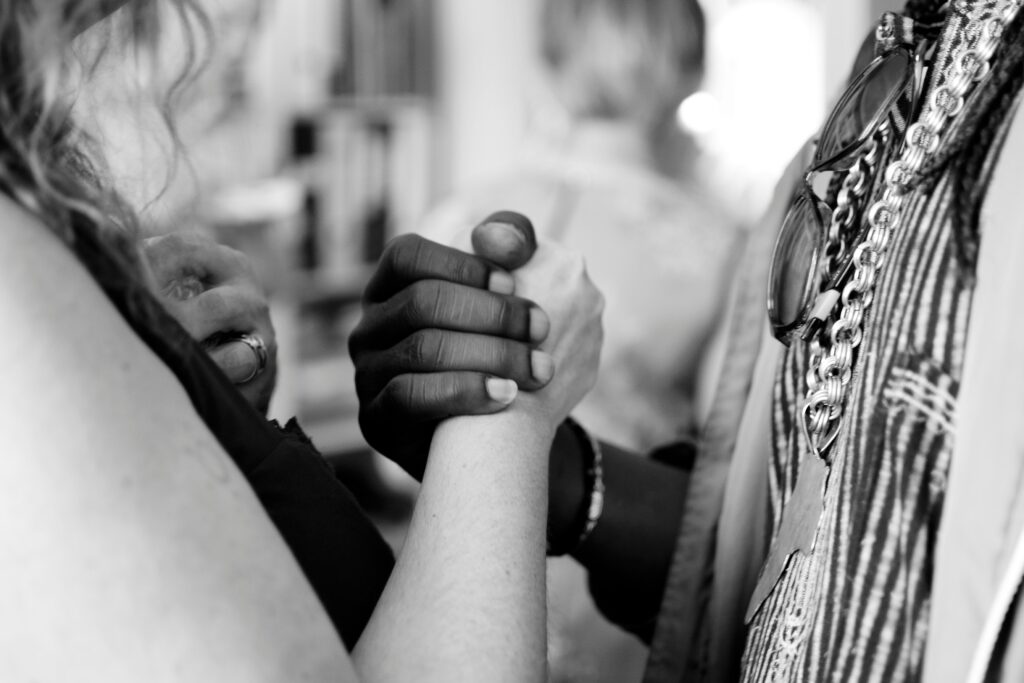 Close-up black and white photo of two people clasping hands in a handshake, symbolizing social progress. One person wears beaded jewelry and the other wears a ring. Blurred background.