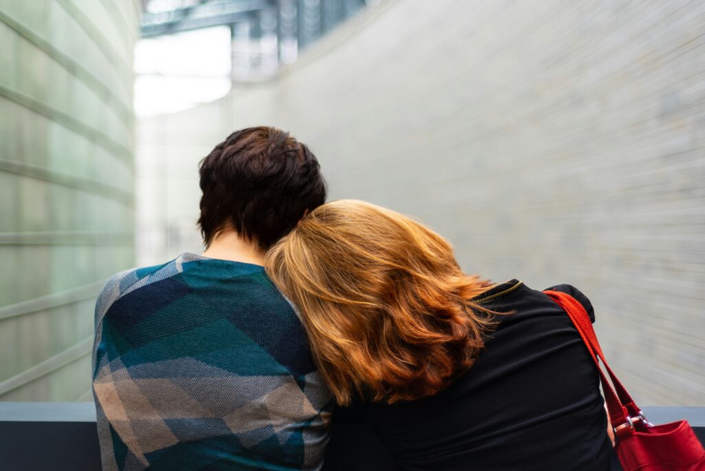 Two people are sitting with their backs to the camera, one resting their head on the other's shoulder, indoors, amidst a modern architectural background, evoking a tender moment that whispers of solace in times of prolonged grief.