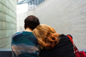 Two people are sitting with their backs to the camera, one resting their head on the other's shoulder, indoors, amidst a modern architectural background, evoking a tender moment that whispers of solace in times of prolonged grief.