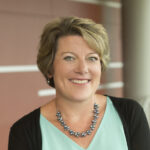 Beth Benatti Kennedy, dressed in a light blue blouse and black cardigan, stands by a railing indoors, smiling.