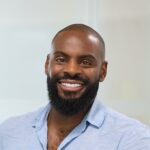 Leon, a man with a beard and shaved head, smiles at the camera in an indoor setting while wearing a light blue button-up shirt.