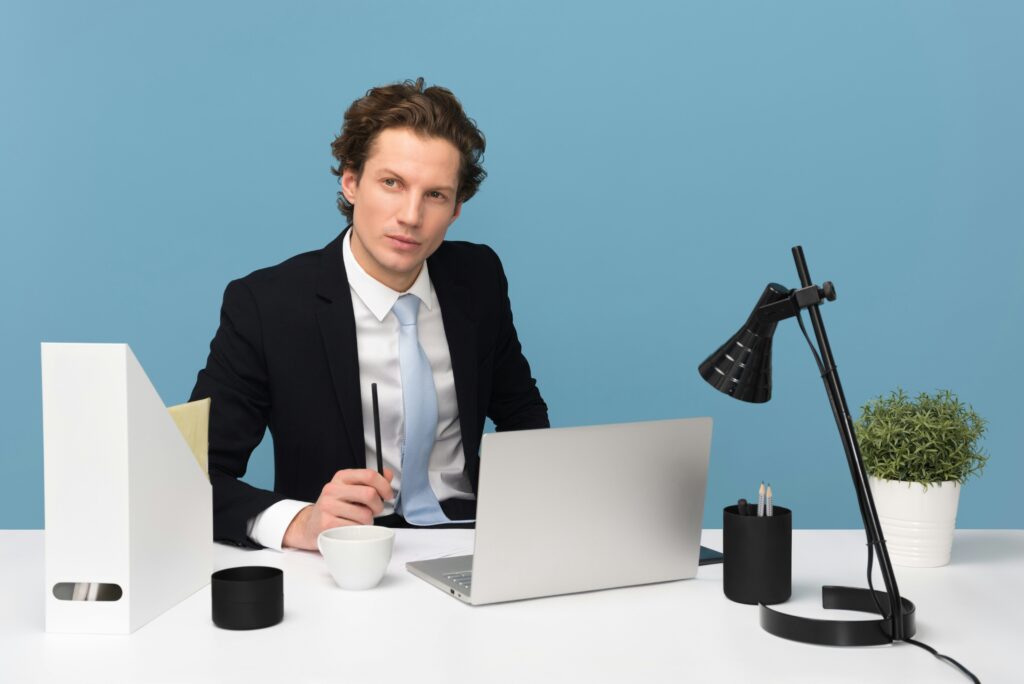 A man in a suit sits at a white desk with a laptop, lamp, plant, and office supplies, holding a pen. He is looking directly at the camera against a blue background, exemplifying the professional environment where neurodivergent workers thrive.