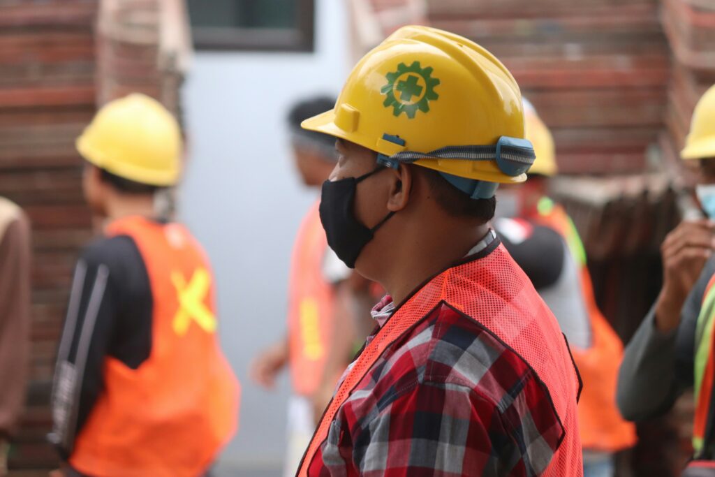 A construction worker wearing a yellow hard hat, face mask, red safety vest, and plaid shirt stands in profile view among other workers also equipped with safety gear.