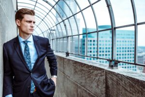 A man in a suit stands on a rooftop next to a metal fence, contemplating the vastness of the city buildings in the background, perhaps reflecting on his role as a poor leader navigating complex urban landscapes.