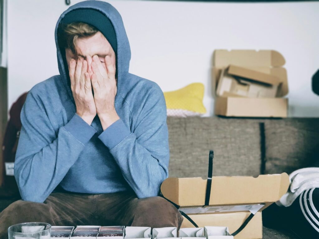 A person in a blue hoodie sits on a couch with their face in their hands, surrounded by open cardboard boxes, anxiously awaiting the relief that payday promises.