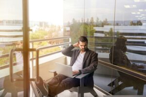 A man in a black suit sits on a balcony with his feet up, exemplifying perfect work-life balance as he taps away on his laptop. Reflections appear on the glass door.