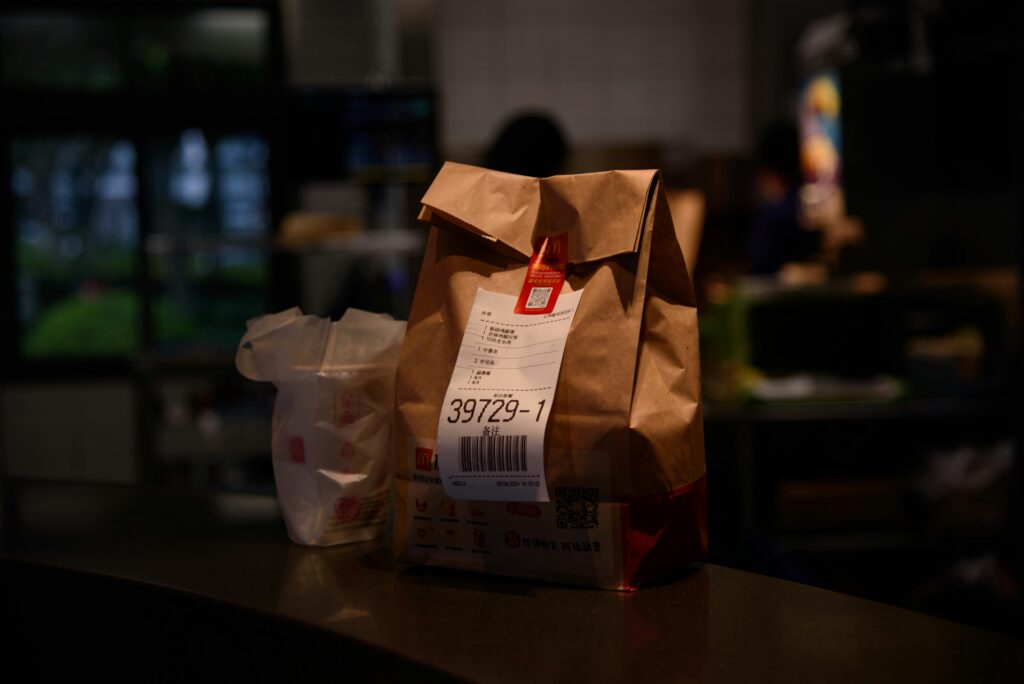 A brown paper bag with a receipt and barcode stands on a counter, next to a white plastic bag, in a dimly lit fast-food restaurant—a stark reminder of modern consumer convenience among whispers of economic chains akin to slavery.