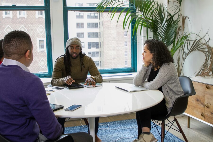 Three people engage in a discussion around a white table in a modern office setting, fostering positive action. Laptops and a smartphone lie within reach, capturing the focus of their collaborative efforts.