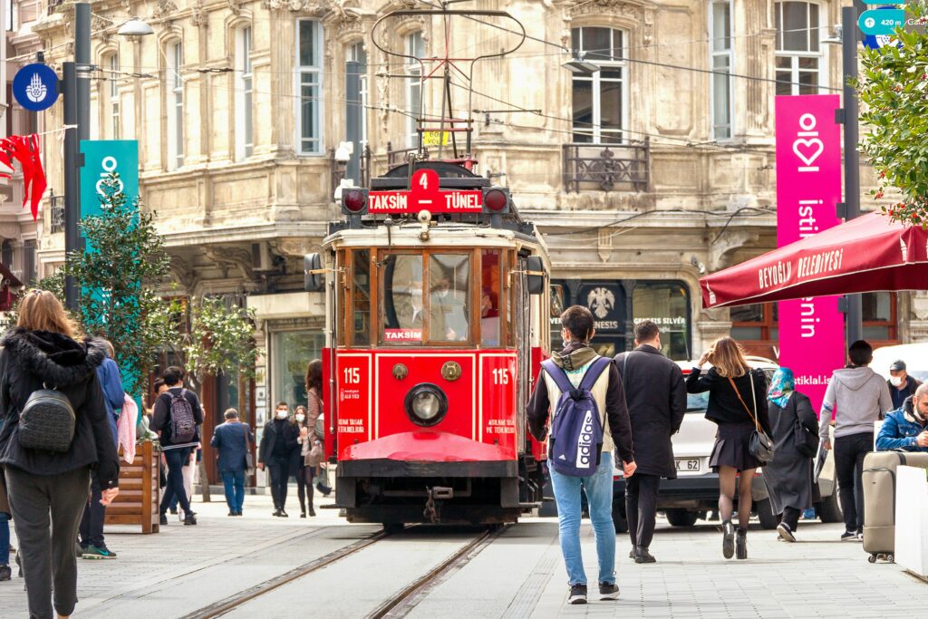 A red tram travels down a bustling street, promoting a sense of wellbeing as pedestrians stroll alongside it. The surrounding buildings showcase traditional architecture.
