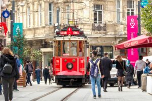 A red tram travels down a bustling street, promoting a sense of wellbeing as pedestrians stroll alongside it. The surrounding buildings showcase traditional architecture.