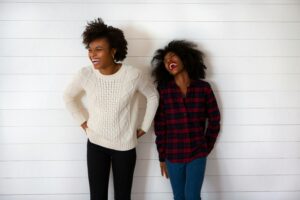 Two people stand side by side against a white wall, both laughing. One wears a white sweater and black pants, the other a red plaid shirt and blue jeans. Their candid joy highlights the importance of fostering mental health in the workplace.