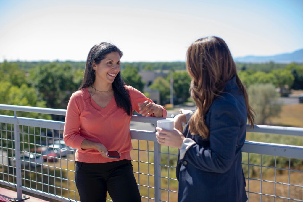 Two people are talking on a balcony, one holding a phone and the other a cup, with increased demand for conversation topics flowing naturally against the backdrop of trees and hills.
