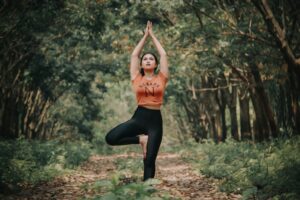 A person embraces wholebeing by practicing tree pose yoga on a forest pathway, surrounded by lush green trees and fallen leaves, wearing an orange top and black leggings.