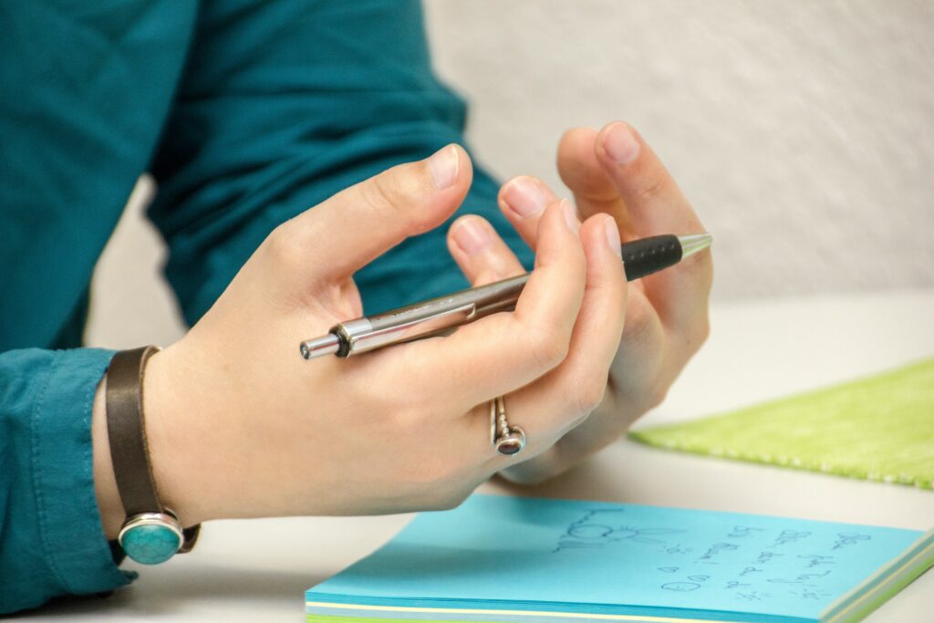 A person wearing a teal shirt and a bracelet holds a pen, gesturing thoughtfully above a blue notepad, perhaps jotting down insights on mental health support.