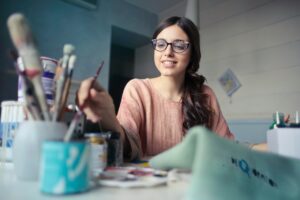 A woman wearing glasses is painting with a brush in her right hand, finding a moment of wellbeing at work. Various art supplies are scattered on the table in front of her, adding color to her creative escape.