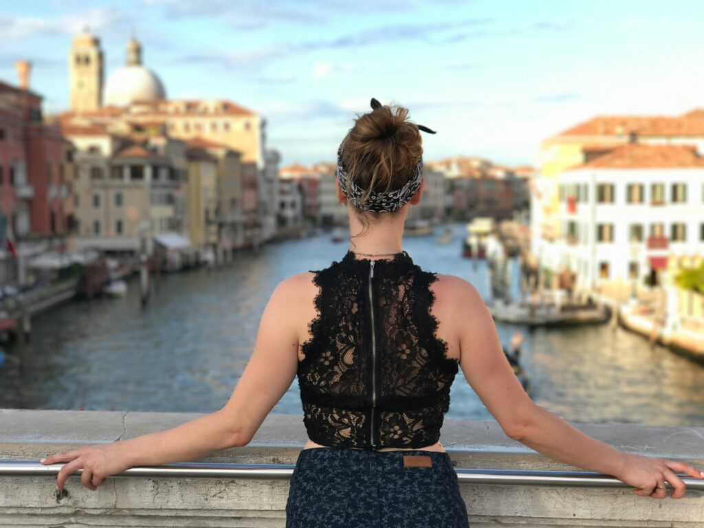 A woman in a bandana and lace top stands on a bridge, empowered and confident, as she overlooks a canal in a city with historic buildings.