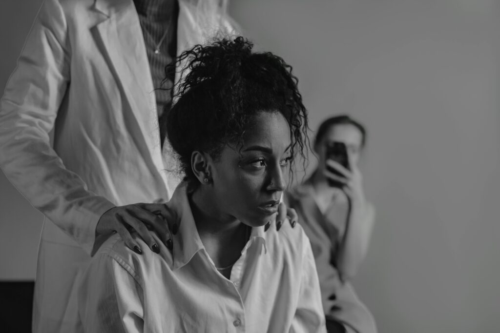 A person in a white shirt sits with a thoughtful expression, their gaze distant, while another offers support with hands gently resting on the shoulders. In the background, someone scrolls through their smartphone, possibly engaging with #MeToo stories.