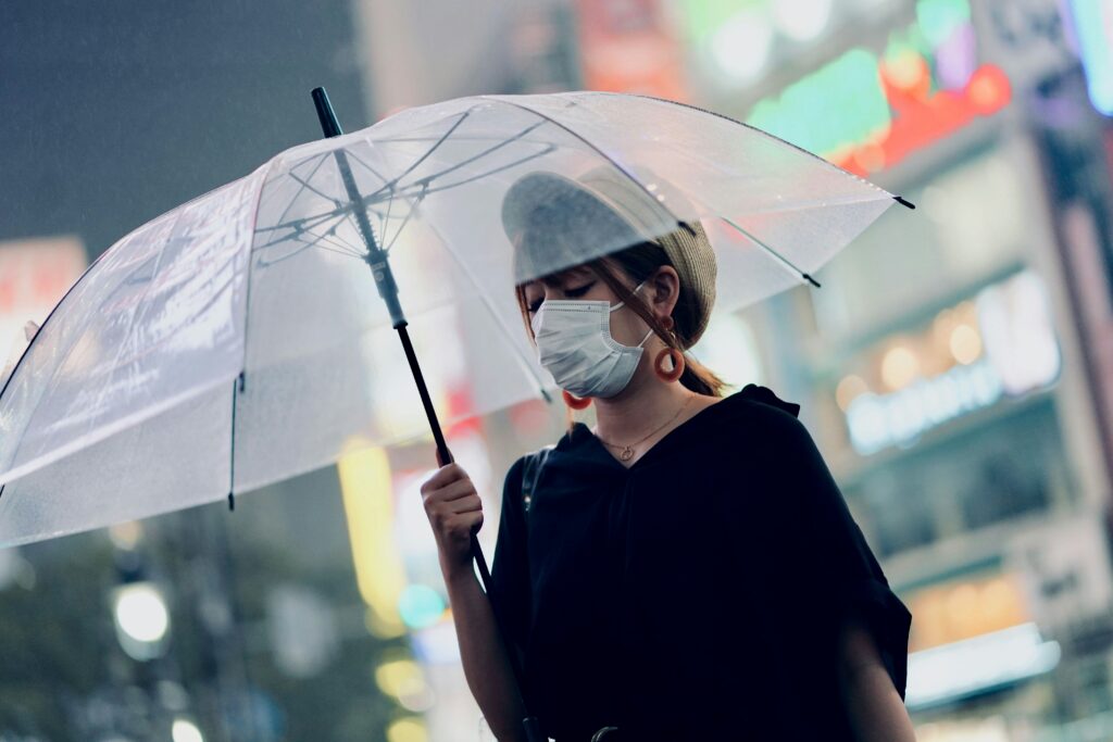 Person holding a transparent umbrella on a rainy night, wearing a face mask to guard against communicable diseases, and a hat. Blurred city lights glow in the background.