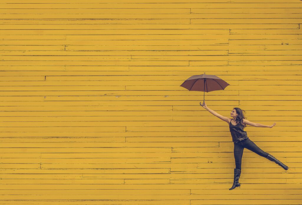 A woman holding an open umbrella is leaping in front of a vibrant yellow wooden wall, embodying the joy and freedom that can come with good mental health. She appears to be floating in mid-air.
