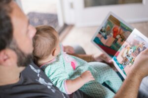 An adult reads a colorful book with their baby and second child while they all sit cozily on the couch.