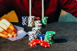 A person in a red hoodie holds playing cards near stacks of poker chips in various colors on a table, a scene that subtly echoes the risks associated with problem gambling.